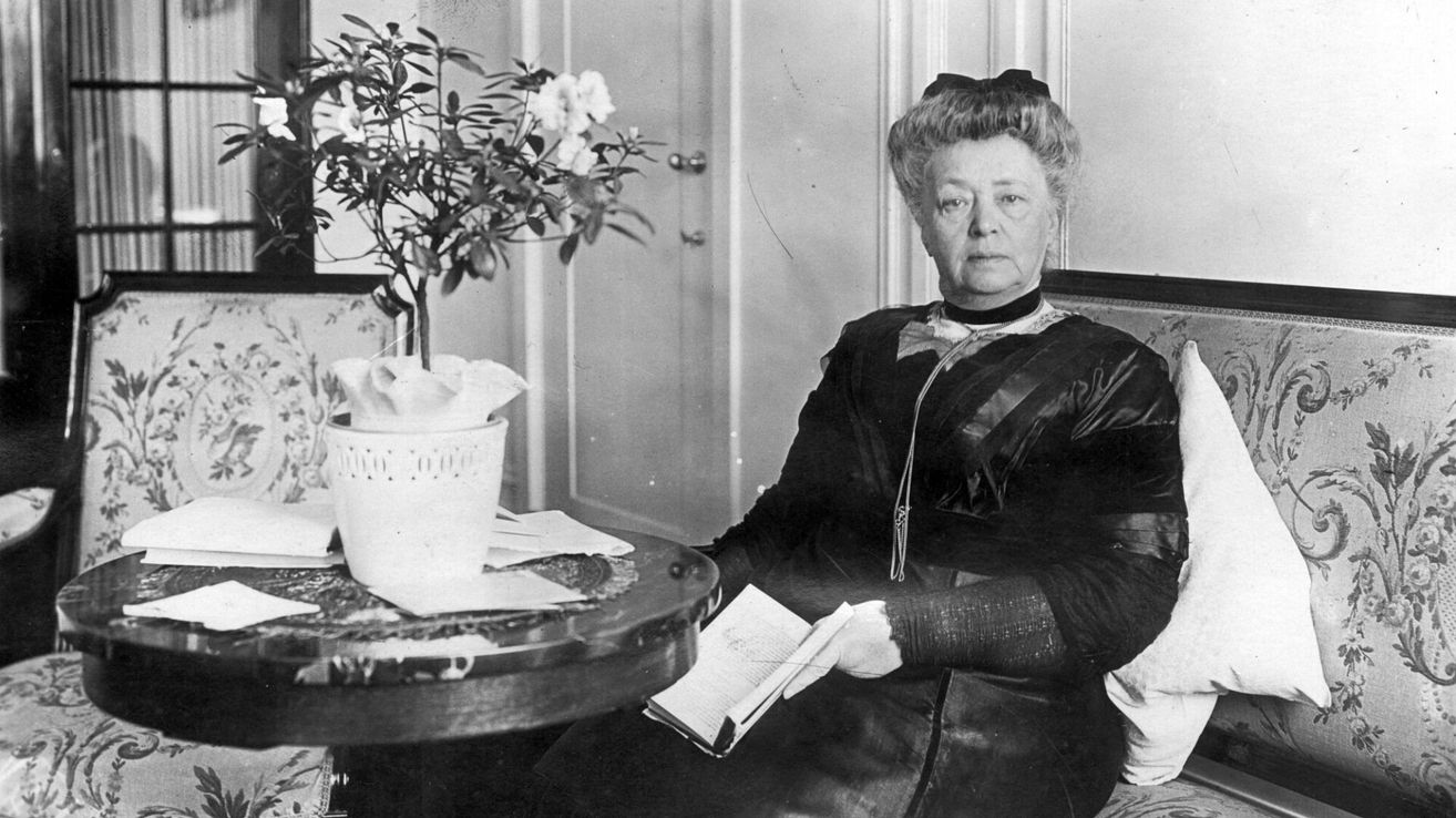 An elderly woman sits in a chair, holding a book, with a potted plant behind her. The room is dimly lit, and she looks serious.