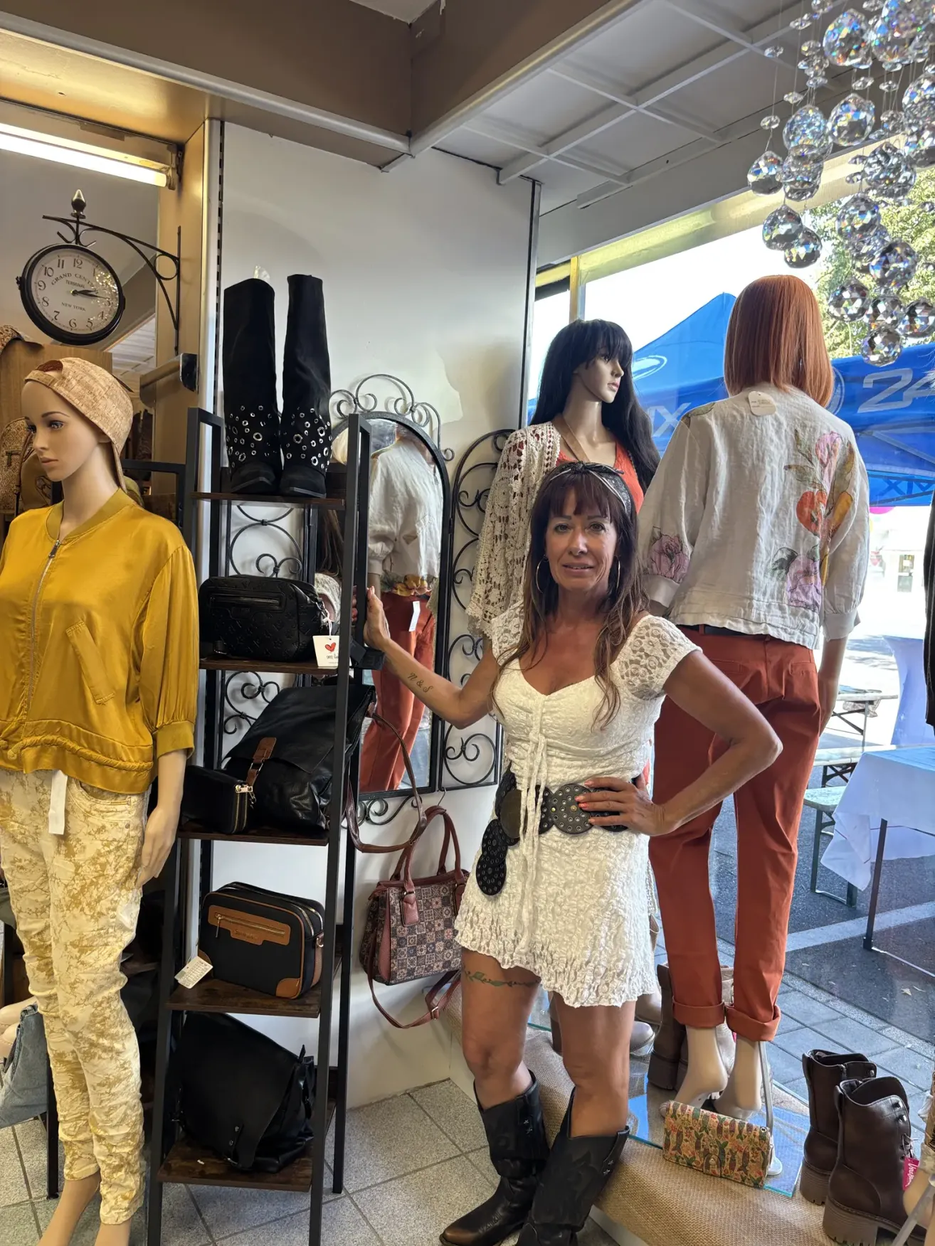 Eine Frau steht in einem Bekleidungsgeschäft, lächelt und posiert mit Mannequins und Kleidungsstücken im Hintergrund.