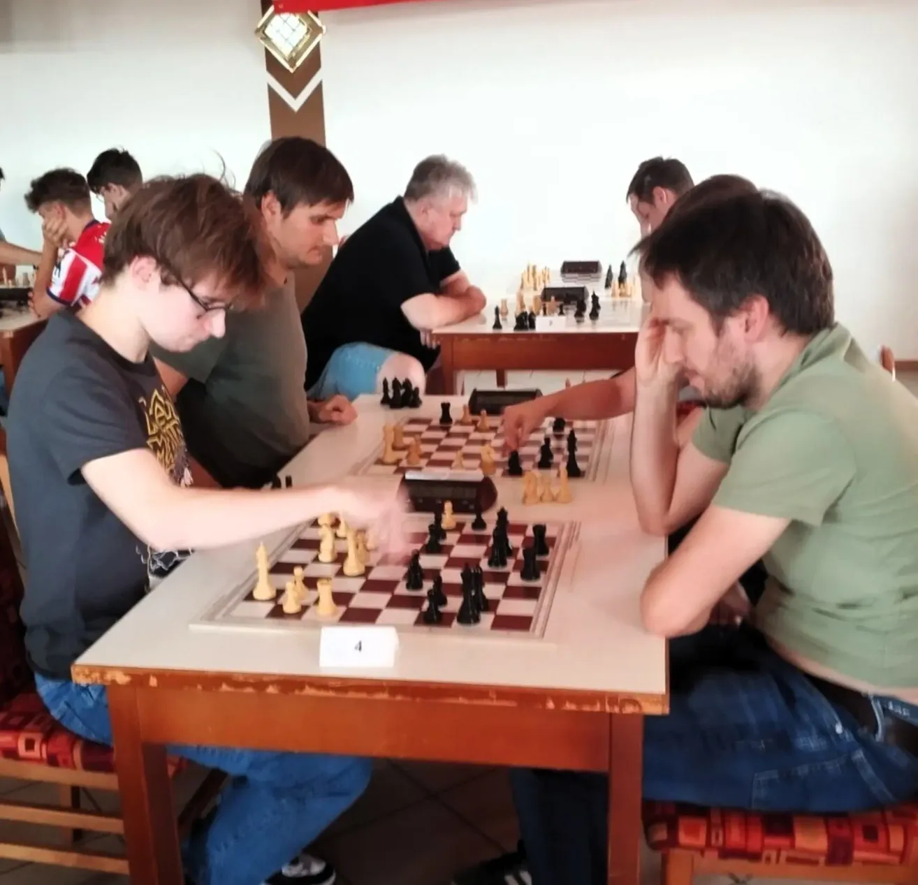 Mehrere Personen spielen Schach in einem Raum. Ein junger Mann mit Brille spielt links, während ein Mann in einem grünen Hemd rechts ist.