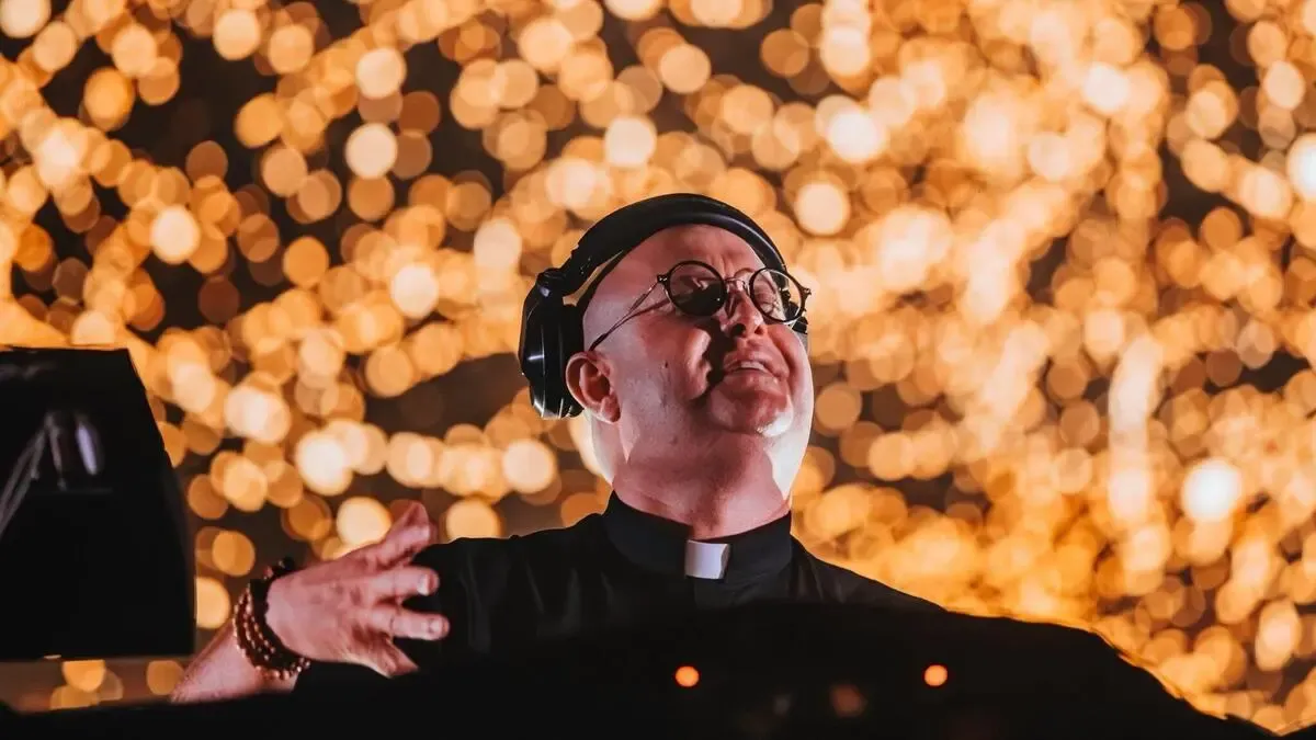 Ein Priester mit Kopfhörern und Brille tritt vor einem verschwommenen Hintergrund von Lichtern auf.