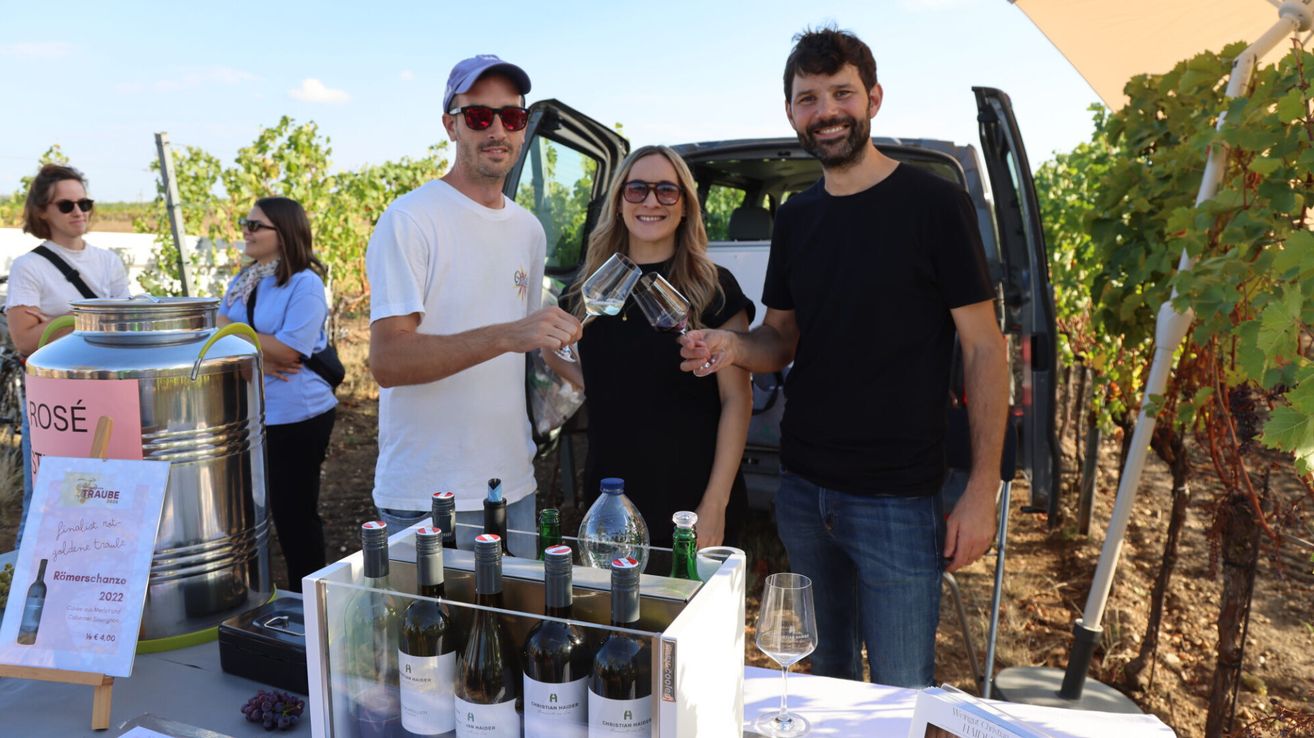 Three people stand in a vineyard, smiling and holding wine glasses. A table with wine bottles and a box is in front of them. A van is parked behind them.