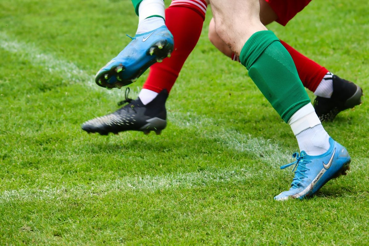 Two soccer players in red and green uniforms are in action, one kicking the ball while the other defends. The image captures a moment of intense competition on a well-maintained grass field.