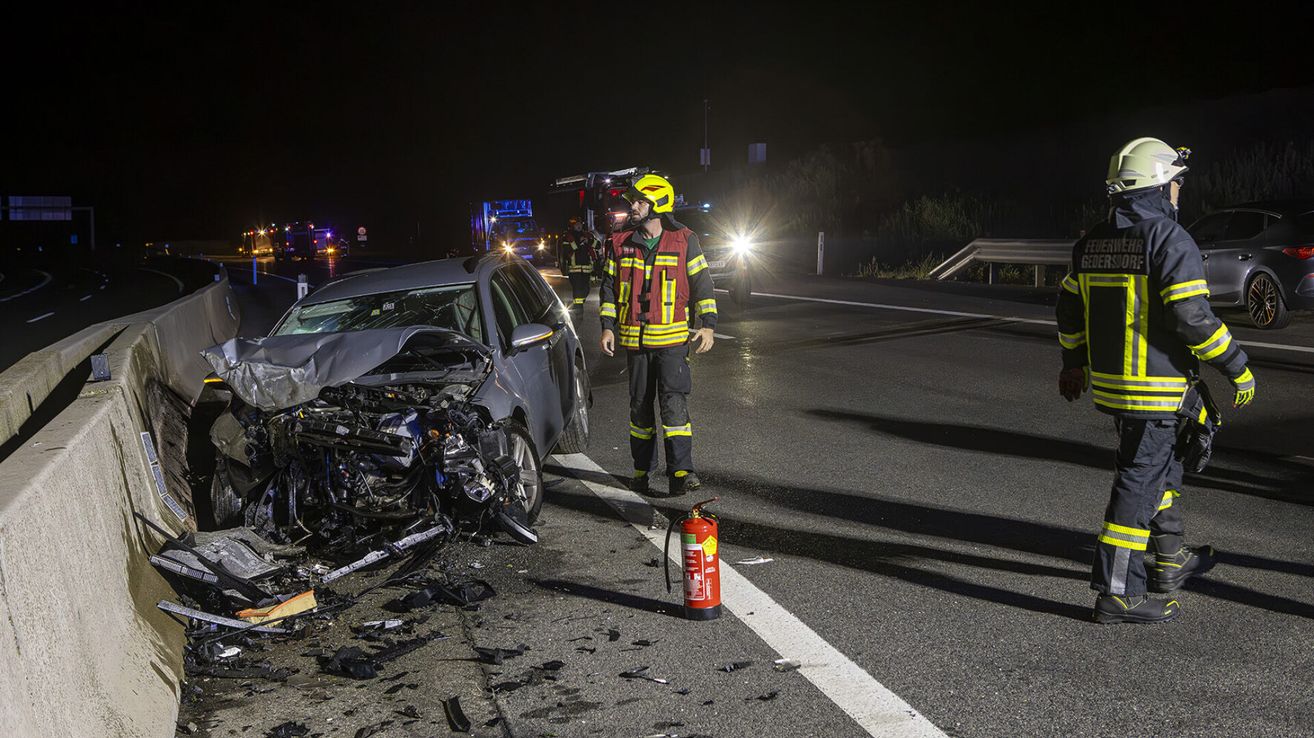 A firefighter in protective gear stands beside a wrecked car on the side of a road at night. The car's front is heavily damaged, with debris scattered around. A fire extinguisher is placed on the road.