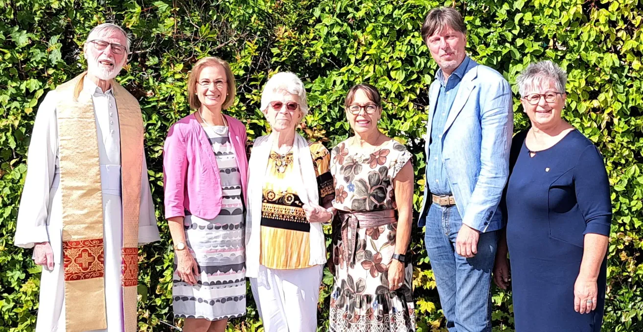 Four people standing in front of a bush, smiling for a photo. The woman on the left wears a pink jacket and a necklace. The woman next to her wears glasses and a white scarf. The woman on the right wears a floral dress and a belt. The man on the right wears a blue shirt and jeans.