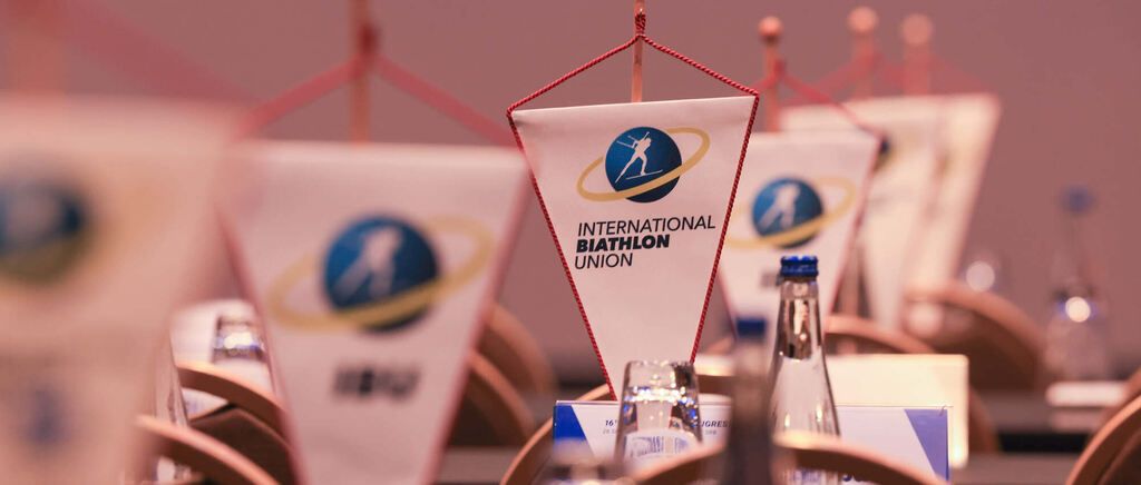 A conference setting with flags of the International Biathlon Union hanging on poles, bottles, and glasses on the table.