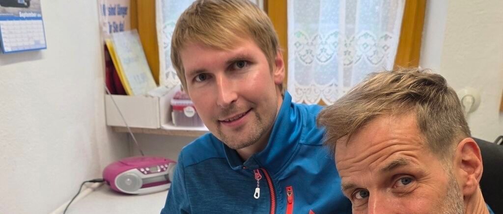 Two men are smiling and posing for a photo in an indoor setting. The man in the foreground is wearing a blue jacket with a zipper, while the man behind him has a serious expression. A pink radio is on a desk in the background, and a shelf with books and other items is visible.