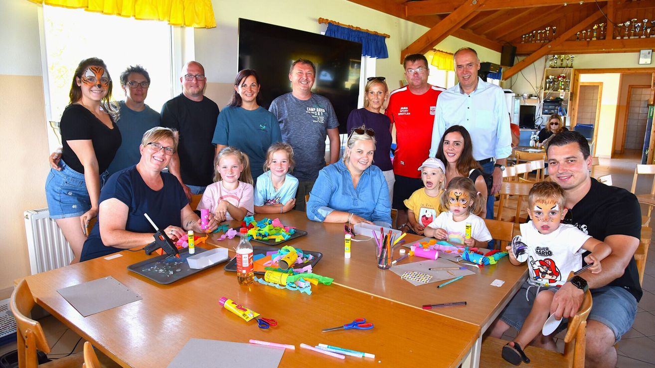 A group of adults and children are gathered around a table, smiling and posing for a photo. The table is filled with art supplies and crafts.