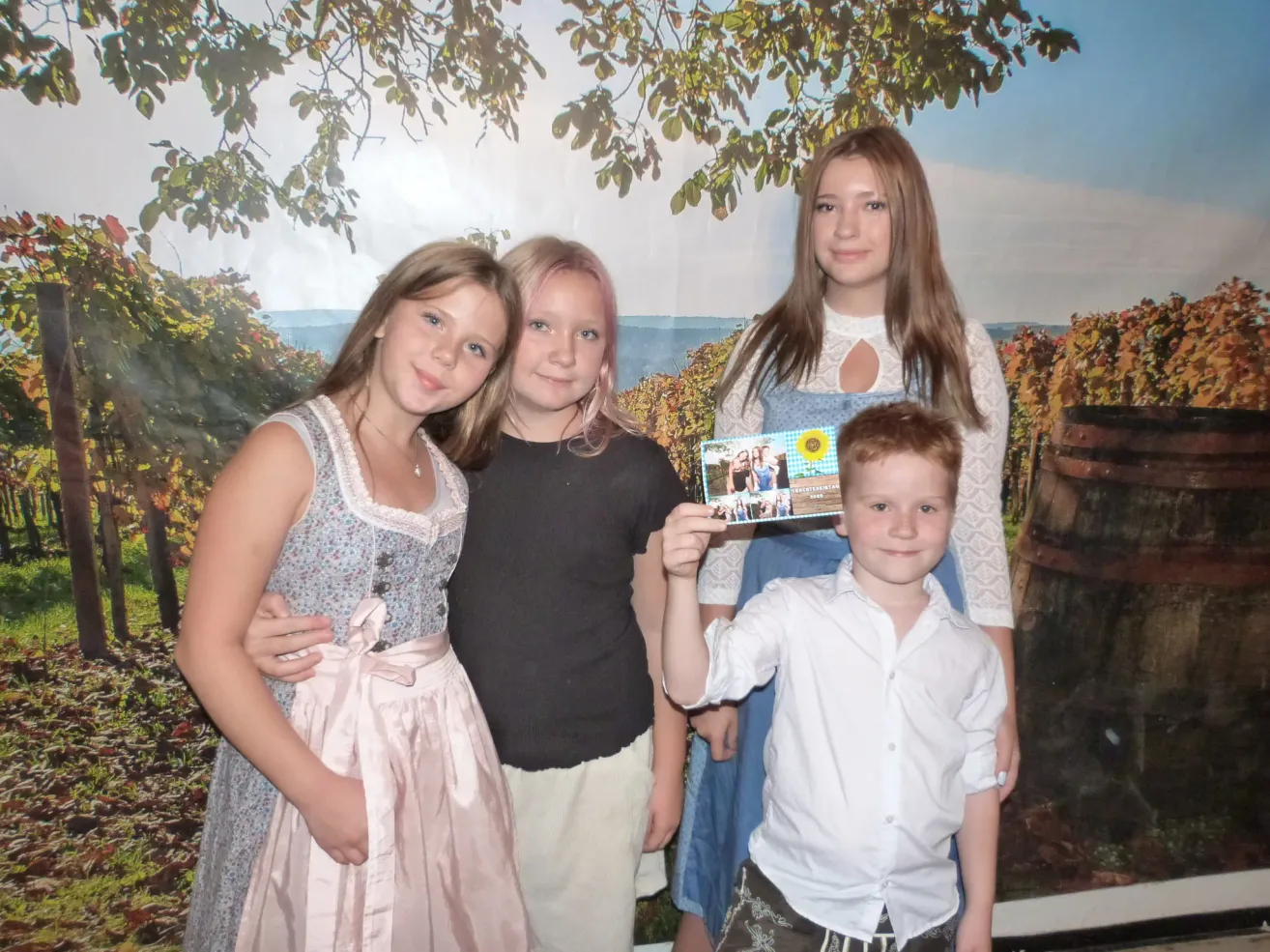 Four children, two girls and two boys, stand in front of a scenic backdrop, smiling and holding a photo. The girl on the left wears a floral dress, while the boy on the right holds a photo.