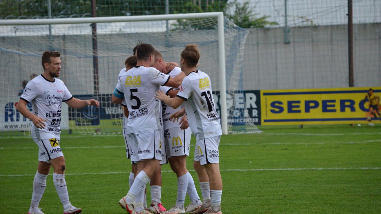 Fußballspieler in weißen Trikots feiern ein Tor in der Nähe des Torpfostens auf einem grünen Feld.