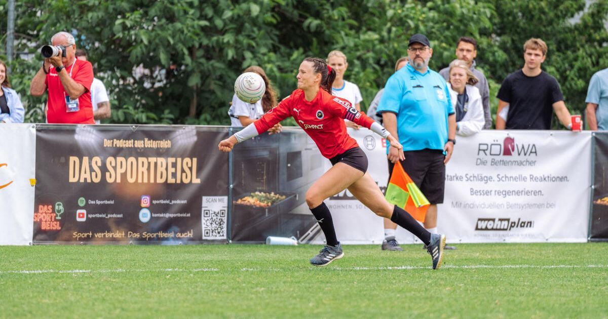 Eine Fußballspielerin in einem roten Trikot ist in der Luft und tritt einen Fußball. Hinter ihr befindet sich ein Banner mit Logo und Text. Zuschauer beobachten das Spiel, darunter ein Mann in einem blauen Hemd.