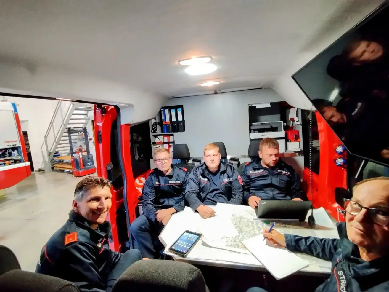 Fünf Feuerwehrleute sitzen um einen Tisch in einem roten Fahrzeug herum und besprechen wahrscheinlich eine Karte und benutzen einen Laptop.