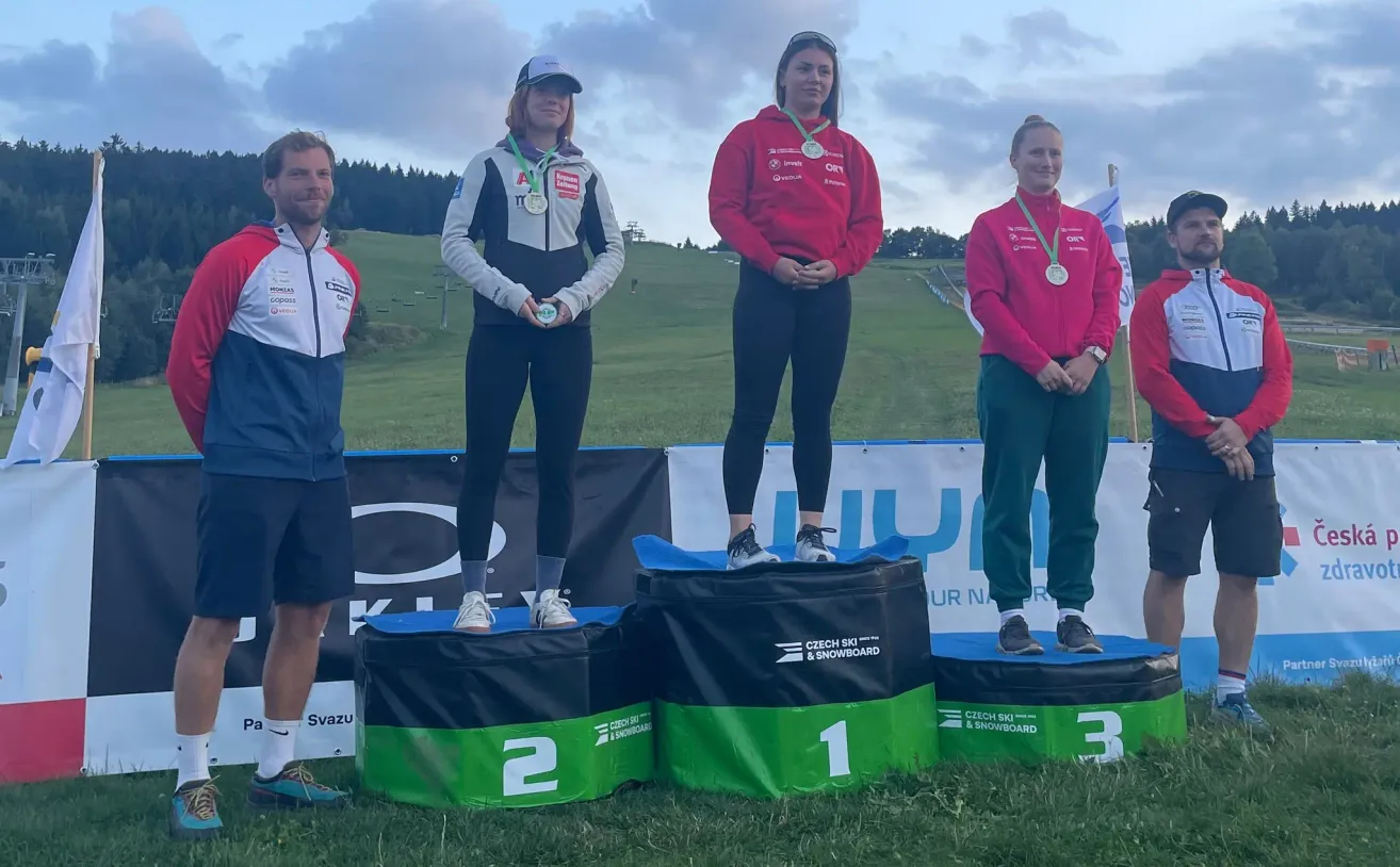 Four athletes stand on a podium with green and black bases, wearing medals. The woman in the middle wears a red jacket and black pants. The woman on the left wears a white jacket with a green lanyard. The woman on the right wears a red jacket and green pants. The man on the left wears a blue and white jacket.