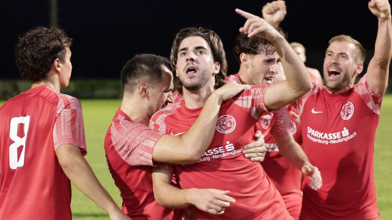 A group of soccer players in red uniforms celebrate a victory on a field at night.