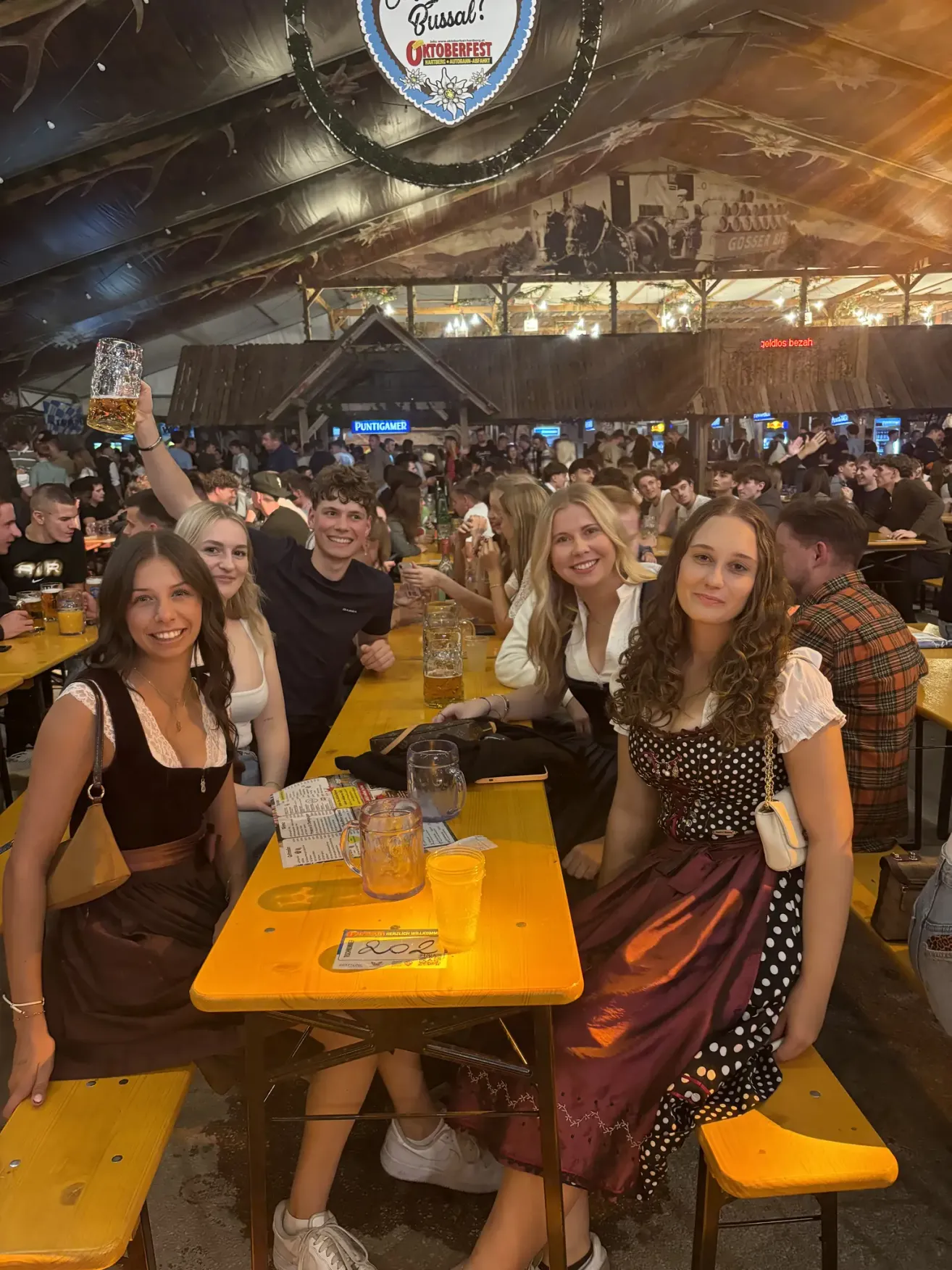 Eine Gruppe von Menschen, darunter Frauen in traditioneller bayerischer Kleidung, sitzen an einem langen Tisch in einem lebhaften Bierzelt und heben Biergläser.