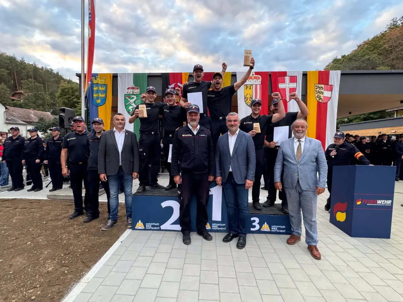 A group of people on a podium with flags in the background, celebrating a victory. Men in uniforms and suits hold trophies and certificates.