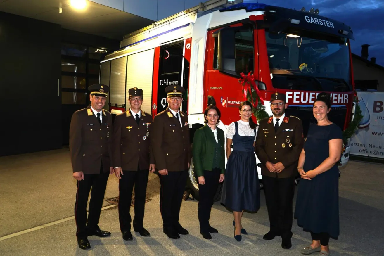 Eine Gruppe von Feuerwehrleuten und Beamten steht vor einem Feuerwehrauto, möglicherweise bei einer Veranstaltung.