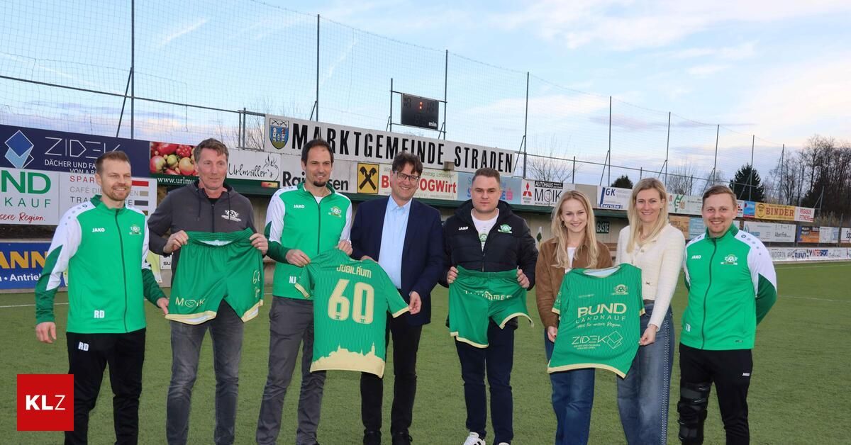 A group of people in green jerseys are posing for a photo on a soccer field. They are holding up their jerseys with numbers and names. Behind them is a fence with banners and a scoreboard.