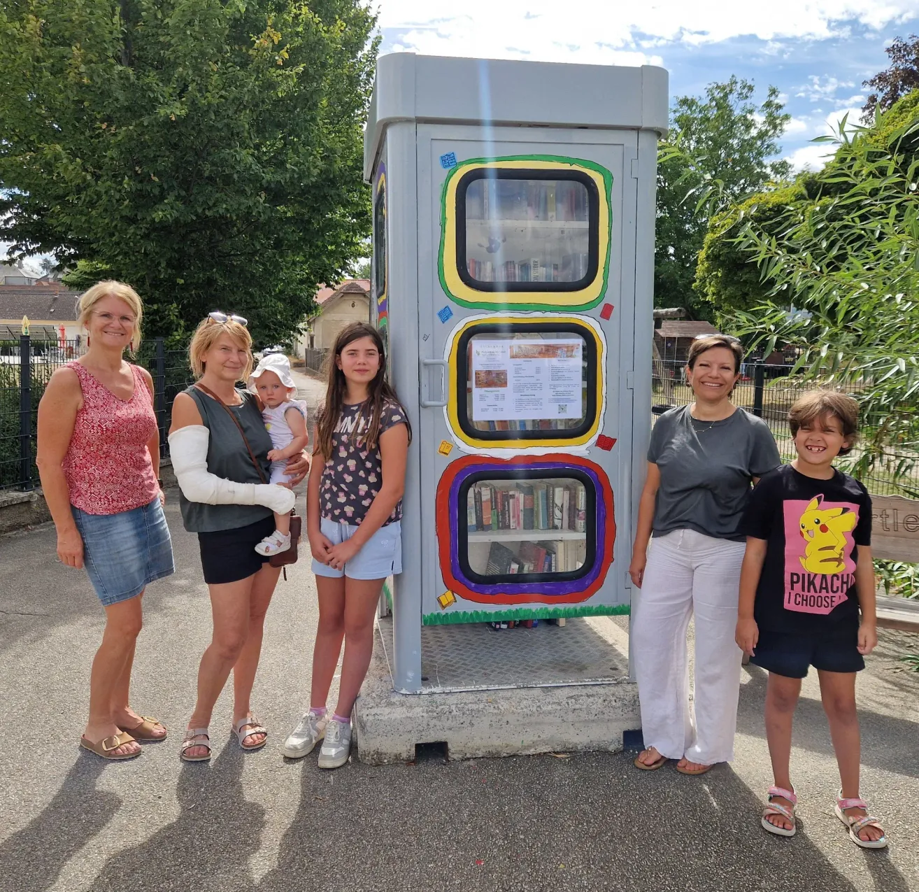 Five women and two children stand in front of a colorful book vending machine in a park.