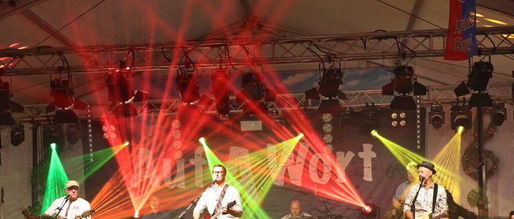 A band performs on stage under vibrant red and green stage lights. The lead singer holds a guitar and sings into a microphone.