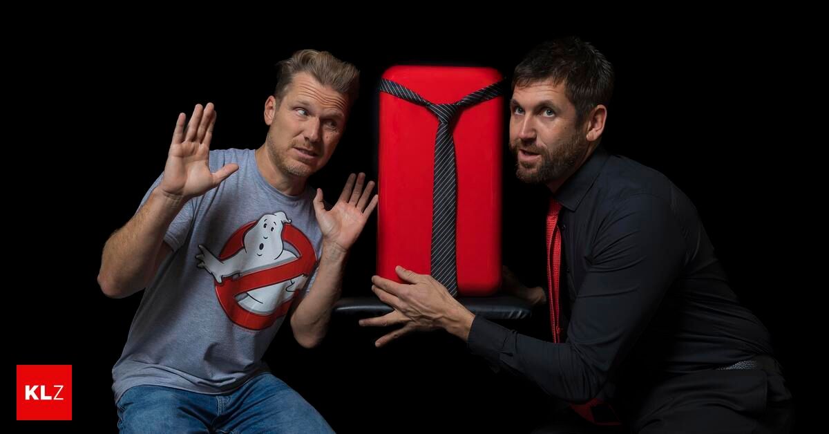 Two men are posing with a red suitcase, one wearing a Ghostbusters t-shirt and the other in a black suit with a red tie.