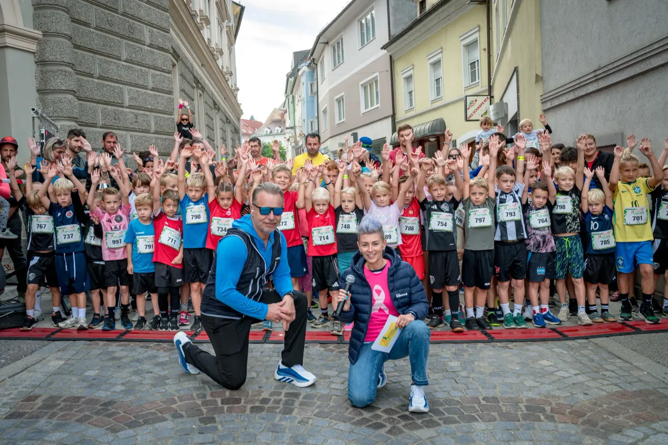 Eine Gruppe von Kindern und Erwachsenen steht in einer Reihe, hebt die Hände und lächelt für ein Foto. Ein Mann und eine Frau knien vor ihnen und halten ein Mikrofon und ein Stück Papier. Die Kinder tragen Hemden mit Nummern.