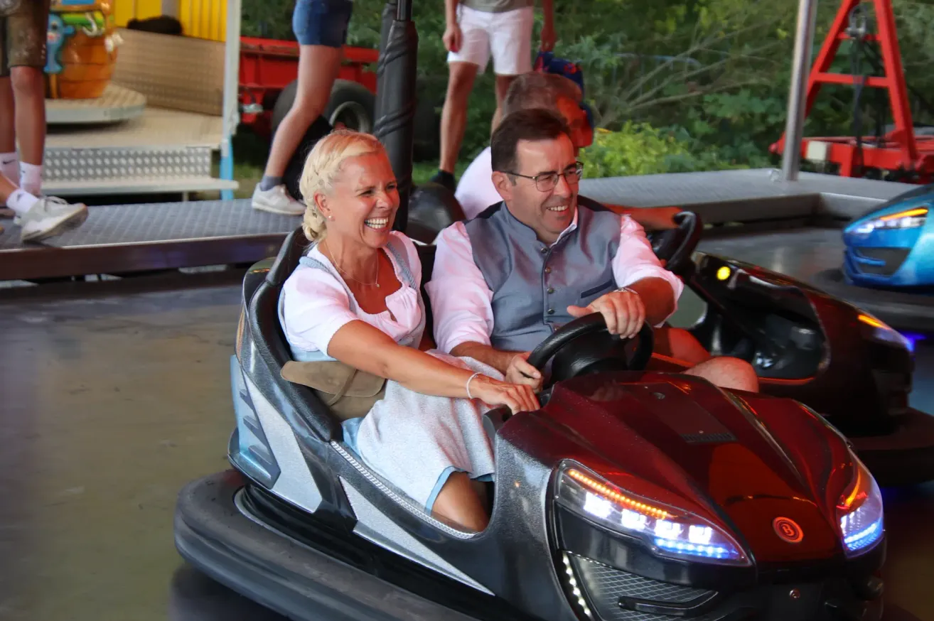 A man and woman are riding a bumper car, smiling and laughing. The woman is holding the steering wheel. Behind them, people are standing near a red truck.