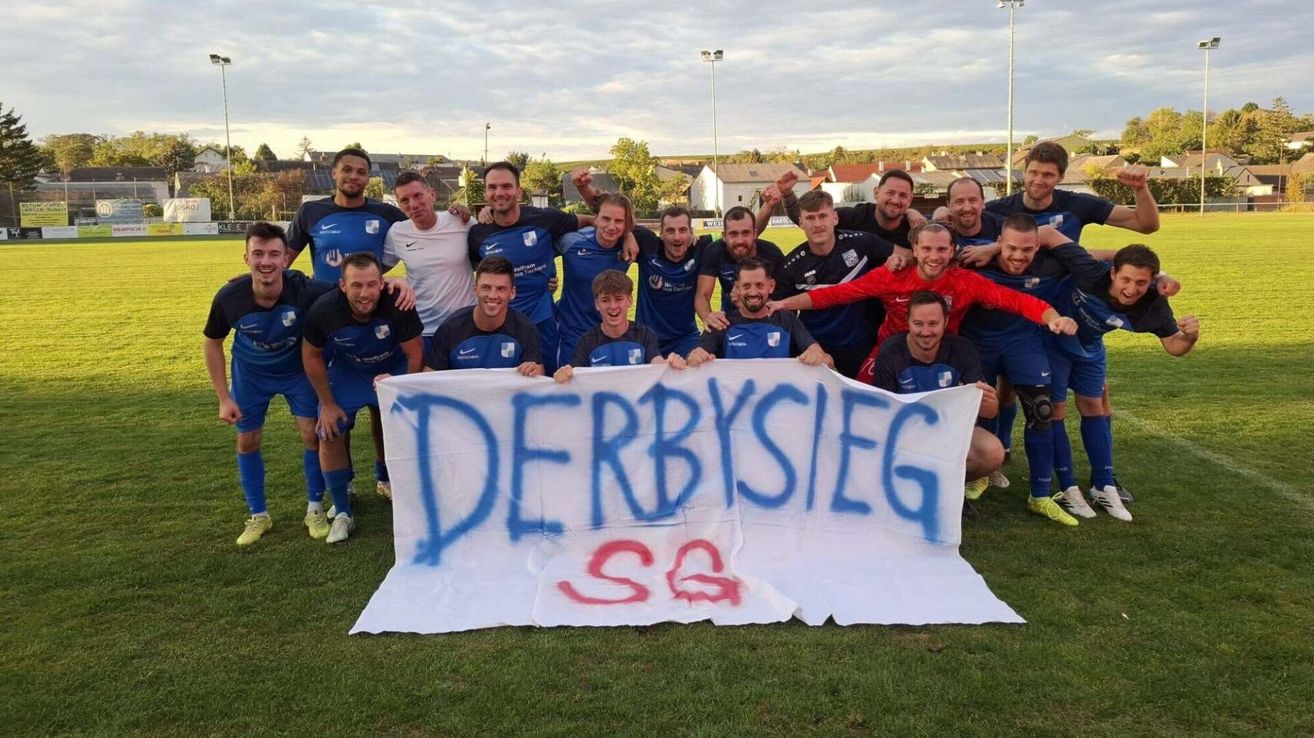 A soccer team celebrates with a banner that reads Derbysieg in blue and red on a grassy field.