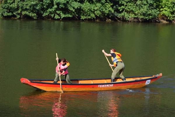 Two people in a canoe paddle down a river, with the word 'Feuerwehr' on the side.