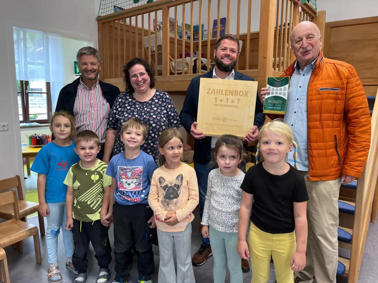 A group of adults and children stand together, smiling, in a room. The adults hold a sign that reads 'Zahlenbach 1+1=?' and a Rotary Club award. The children wear casual clothing and some have glasses. The room has wooden furniture and a wooden railing.