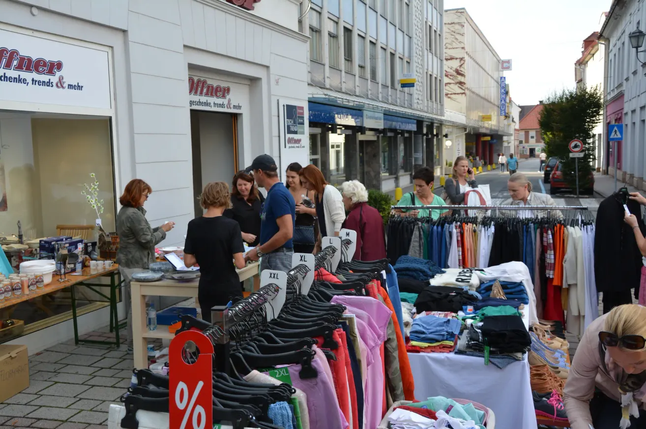 Ein Straßenmarkt mit verschiedenen Ständen, die Kleidung verkaufen. Leute stöbern durch die Kleider an Kleiderbügeln und Tischen. Die Stände sind vor einem Gebäude mit dem Schriftzug 'Offner' angeordnet.