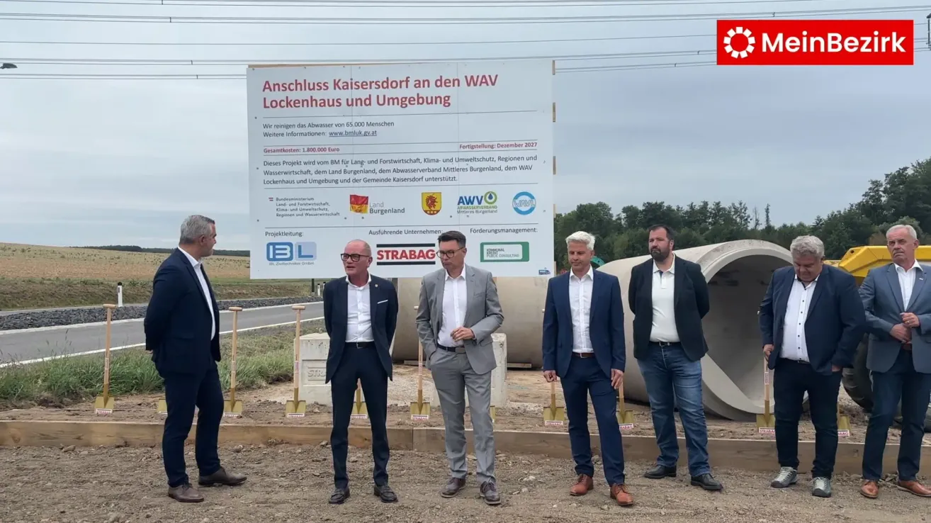 Five men in suits stand in front of a large sign that reads 'Anschluss Kaisersdorf an den WAV Lockenhaus und Umgebung'. They are holding shovels and appear to be at a groundbreaking ceremony. Behind them, there are large pipes and a backdrop of trees.