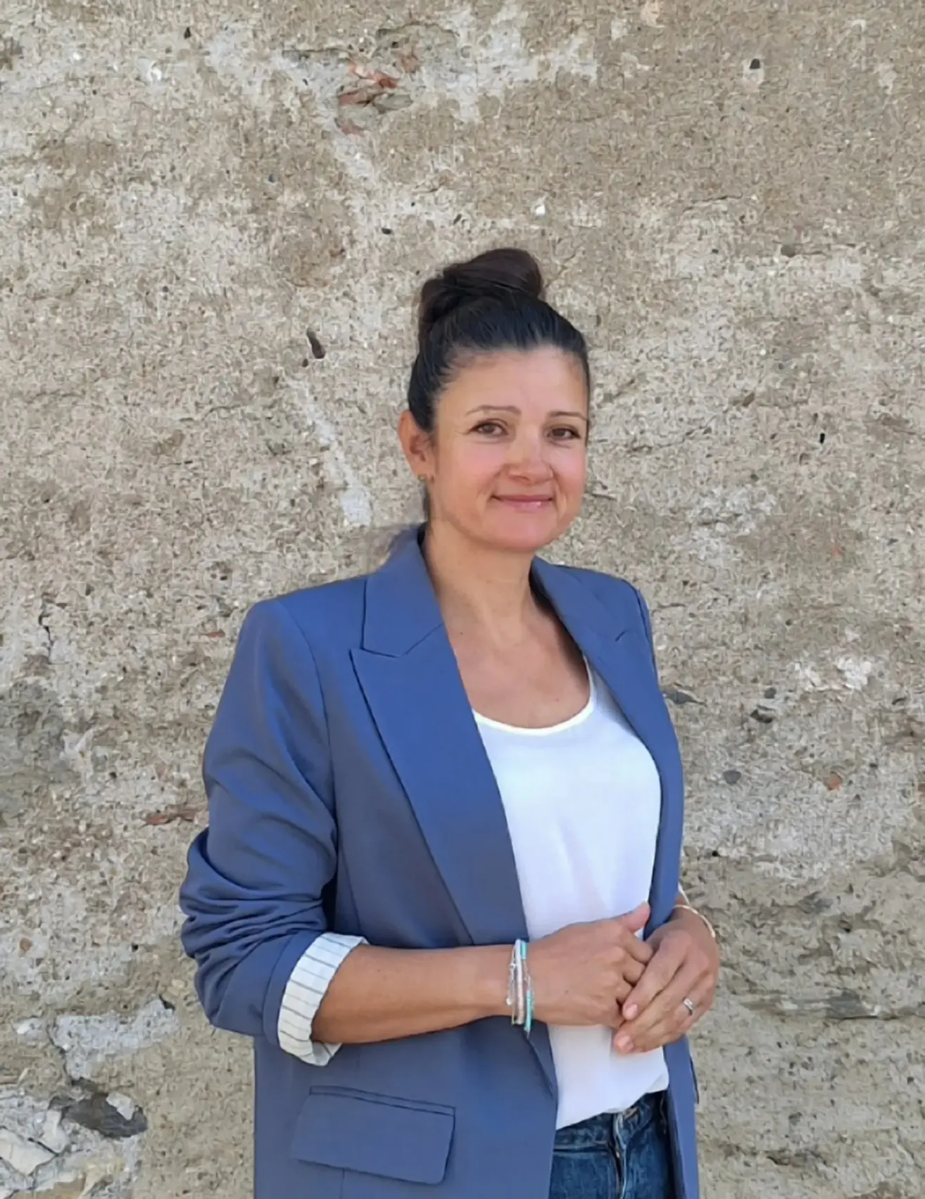 Eine Frau mit Duttfrisur und blauem Blazer steht vor einer strukturierten Wand, lächelt und posiert für ein Foto.