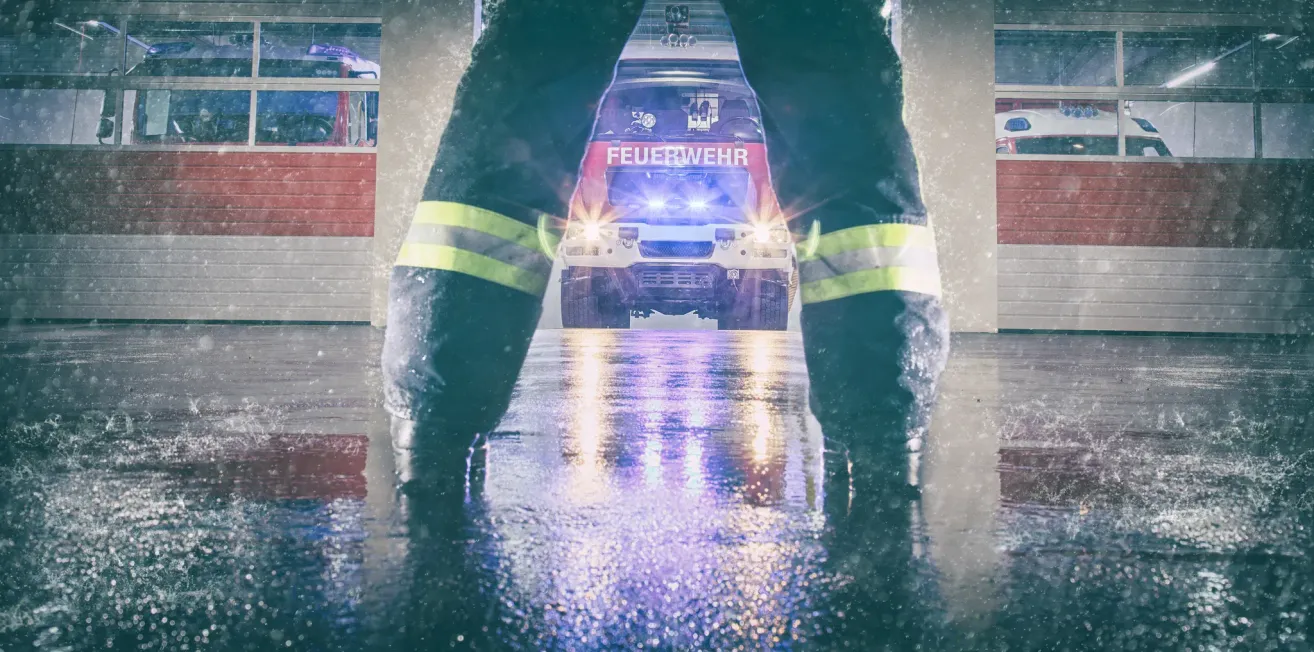 Ein Feuerwehrmann in voller Ausrüstung steht in einem nassen Bereich, mit einem Feuerwehrauto im Hintergrund. Das Auto hat das Wort 'Feuerwehr' darauf.