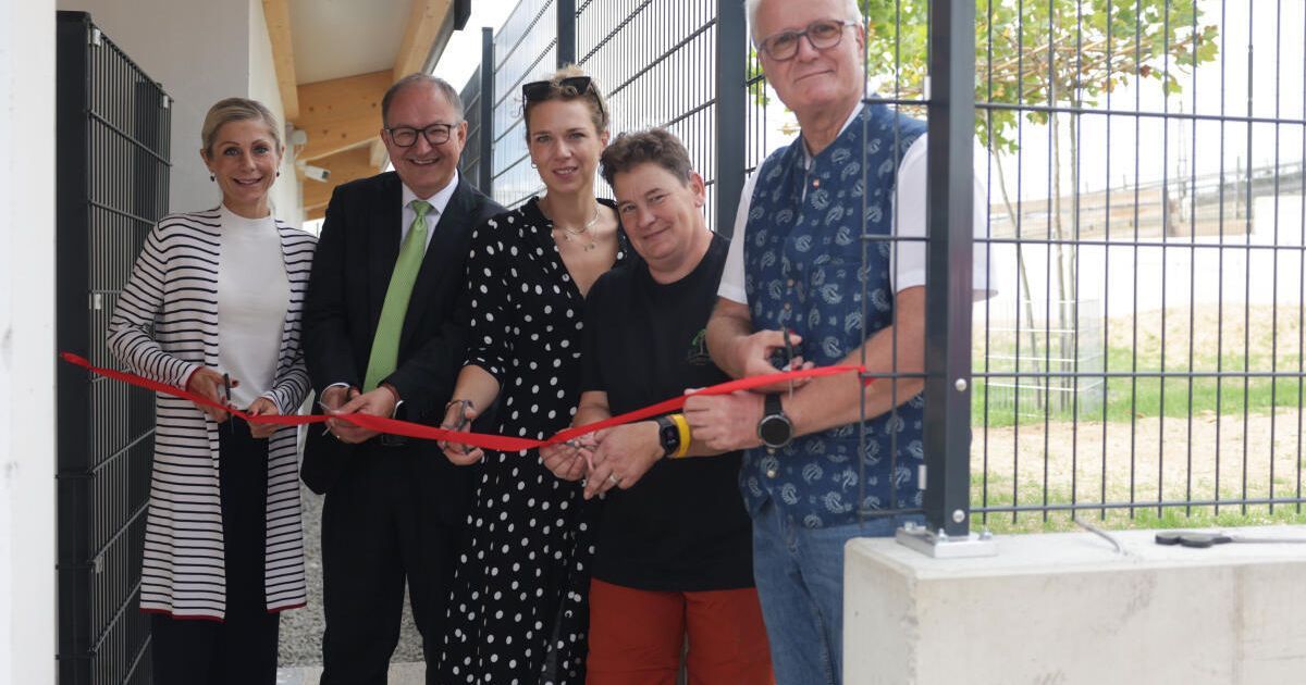 Eine Gruppe von Menschen steht hinter einem Zaun, hält ein rotes Band und lächelt für ein Foto. Sie feiern die Eröffnung einer neuen Einrichtung.