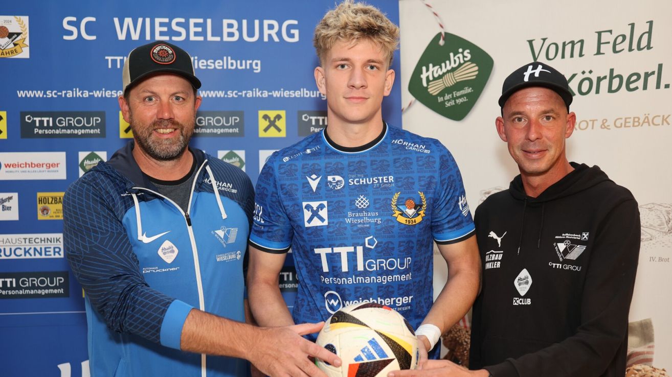 Ein junger Fußballspieler in einem blauen Trikot hält einen Fußball und wird von zwei Männern in blauen und schwarzen Trikots flankiert. Der Hintergrund zeigt verschiedene Logos und Texte, darunter 'Wieselburg' und 'Haubis'.