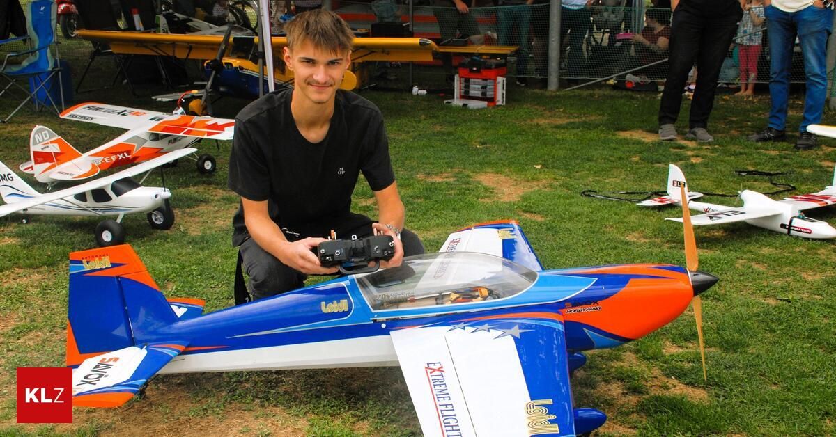 Ein junger Mann sitzt auf dem Rasen und hält eine Fernbedienung für ein blau-oranges Modellflugzeug.