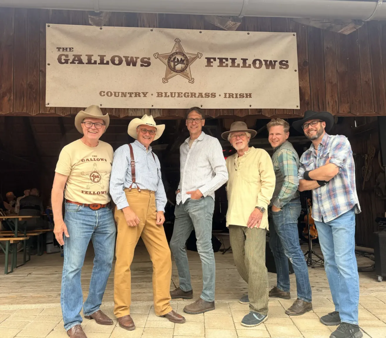 Six men stand in front of a wooden building with a banner reading 'The Gallows Fellows'. They are all wearing hats and smiling for the camera.
