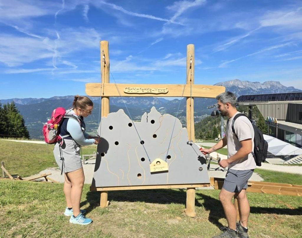 Two hikers, a man and a woman, are standing in front of a wooden structure with a gray board and holes, possibly engaged in a game or activity. They are both wearing backpacks and sneakers. The background shows mountains and a clear blue sky.