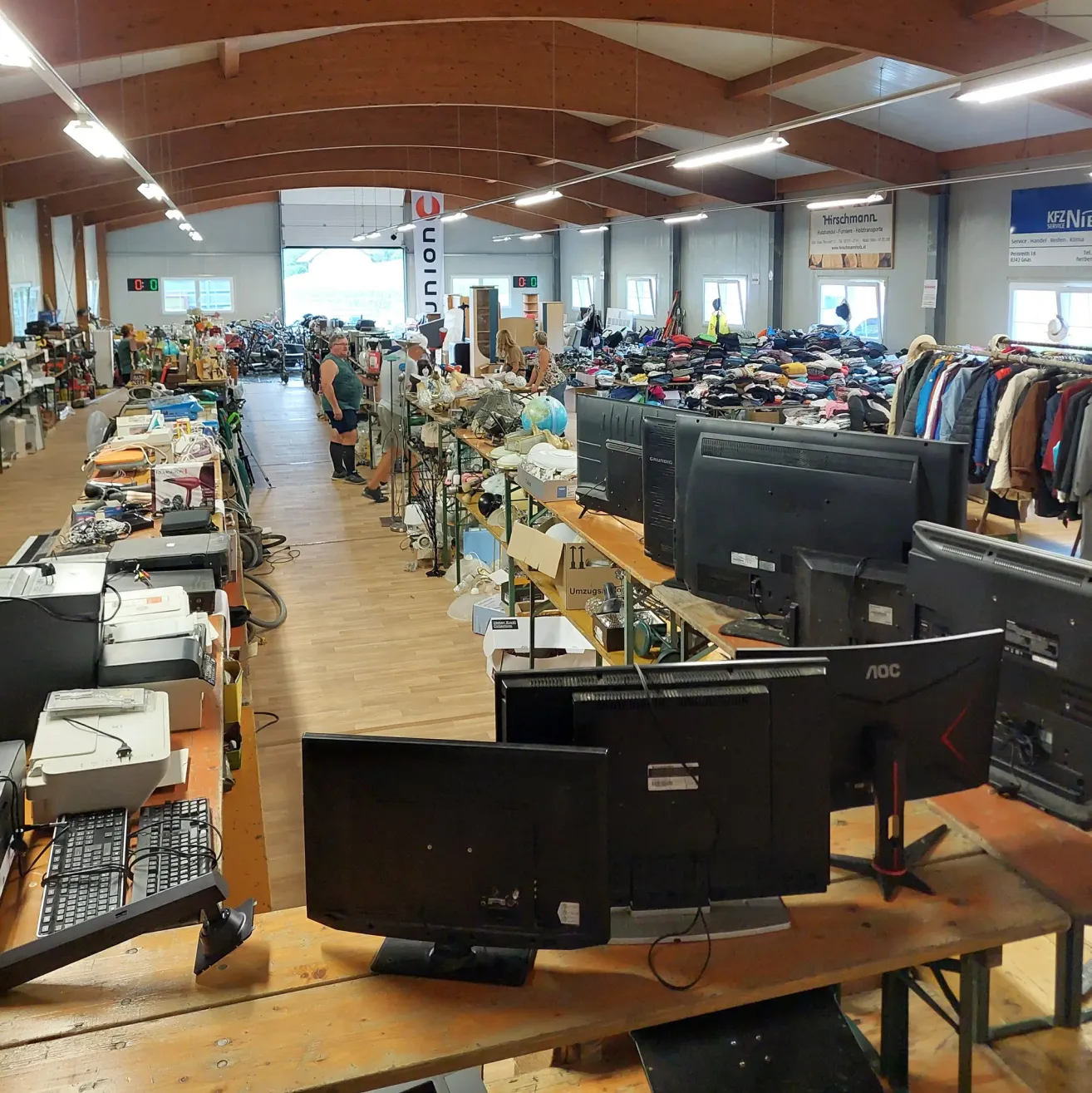 A large indoor space filled with various items for sale, including electronics, clothing, and household goods. People are browsing through the items.