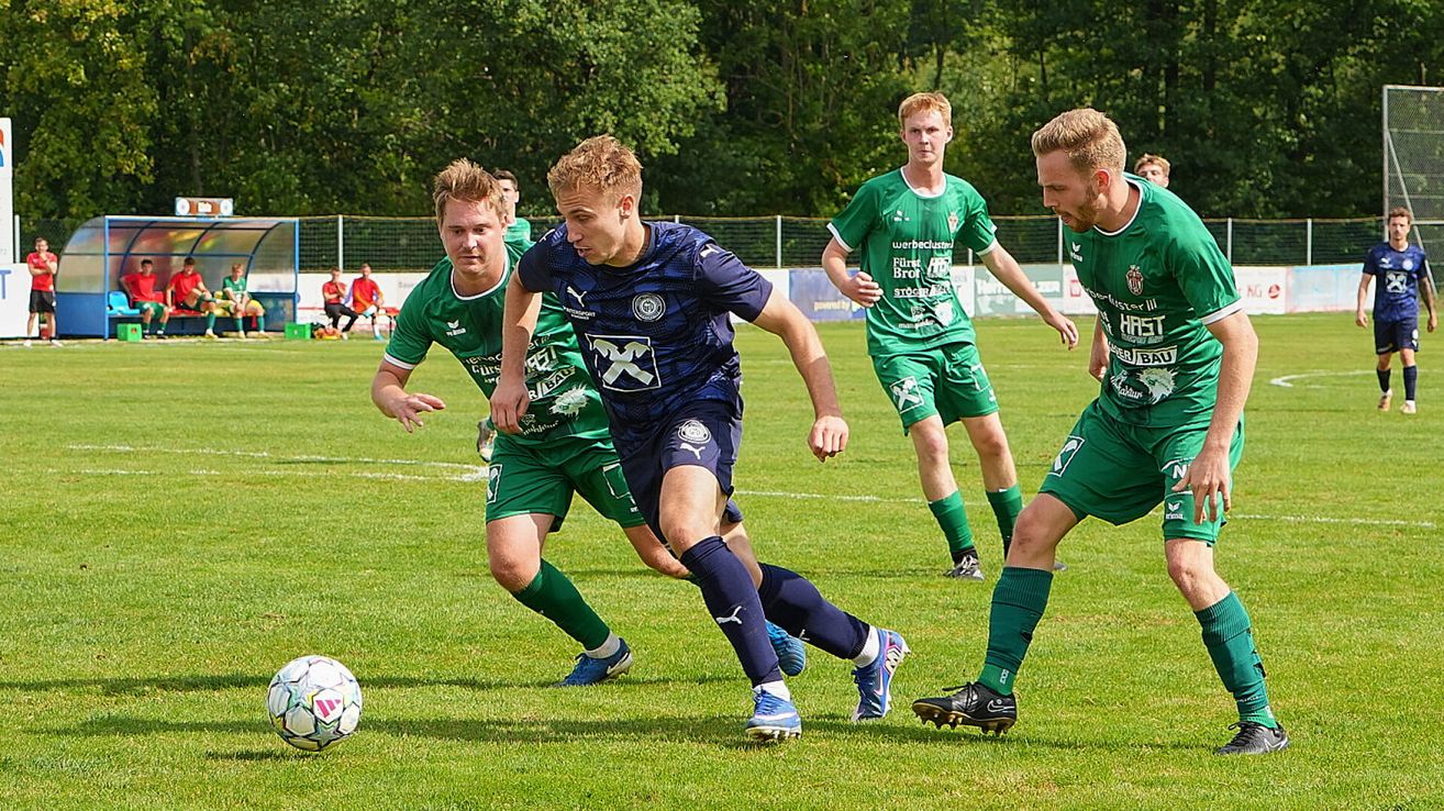 Eine Gruppe von Fußballspielern in grünen und blauen Trikots spielt auf einem Rasenfeld. Ein Spieler in Blau läuft auf den Ball zu, während andere ihn verfolgen.