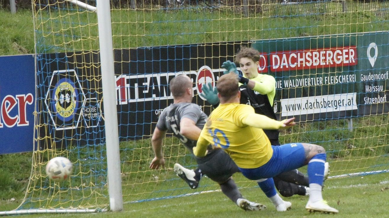 Fußballspieler in Aktion, einer verteidigt das Tor, während der andere versucht zu treffen. Der Torwart trägt grüne Handschuhe.