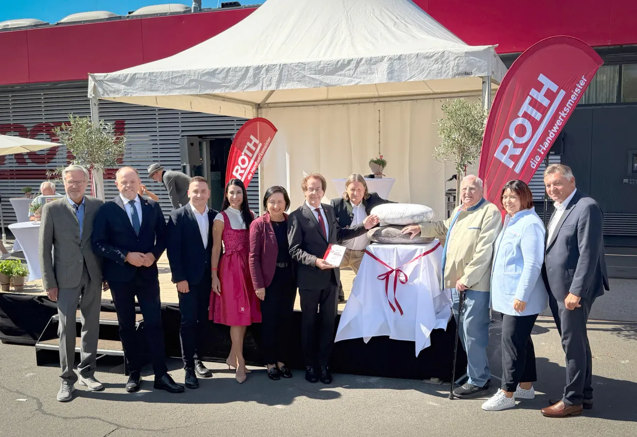 A group of people stand under a tent, smiling for a photo. A man in the center holds a book. A table with a red ribbon is in front of them.