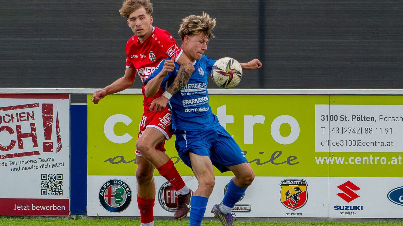 Two soccer players, one in red and the other in blue, are fiercely competing for the ball. The player in blue is about to kick the ball while the player in red is trying to block him. Behind them is a green banner with logos and text.