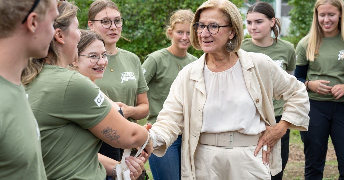 Eine ältere Frau mit Brille lächelt und schüttelt die Hand einer Gruppe junger Frauen, die grüne T-Shirts mit einem Logo tragen.
