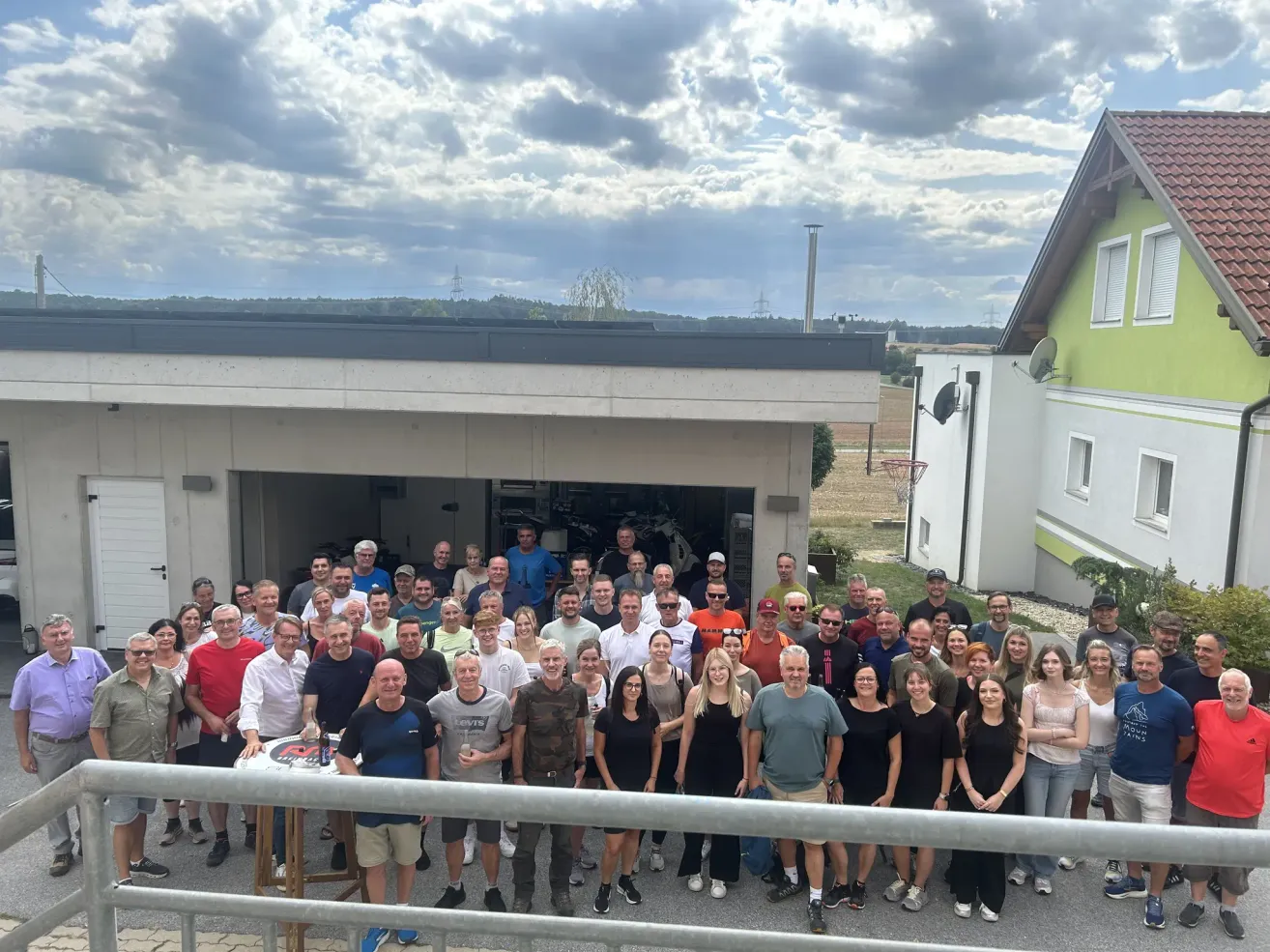 Eine große Gruppe von Menschen, wahrscheinlich ein Team oder eine Familie, posiert für ein Foto vor einem Gebäude mit einem bewölkten Himmel im Hintergrund.