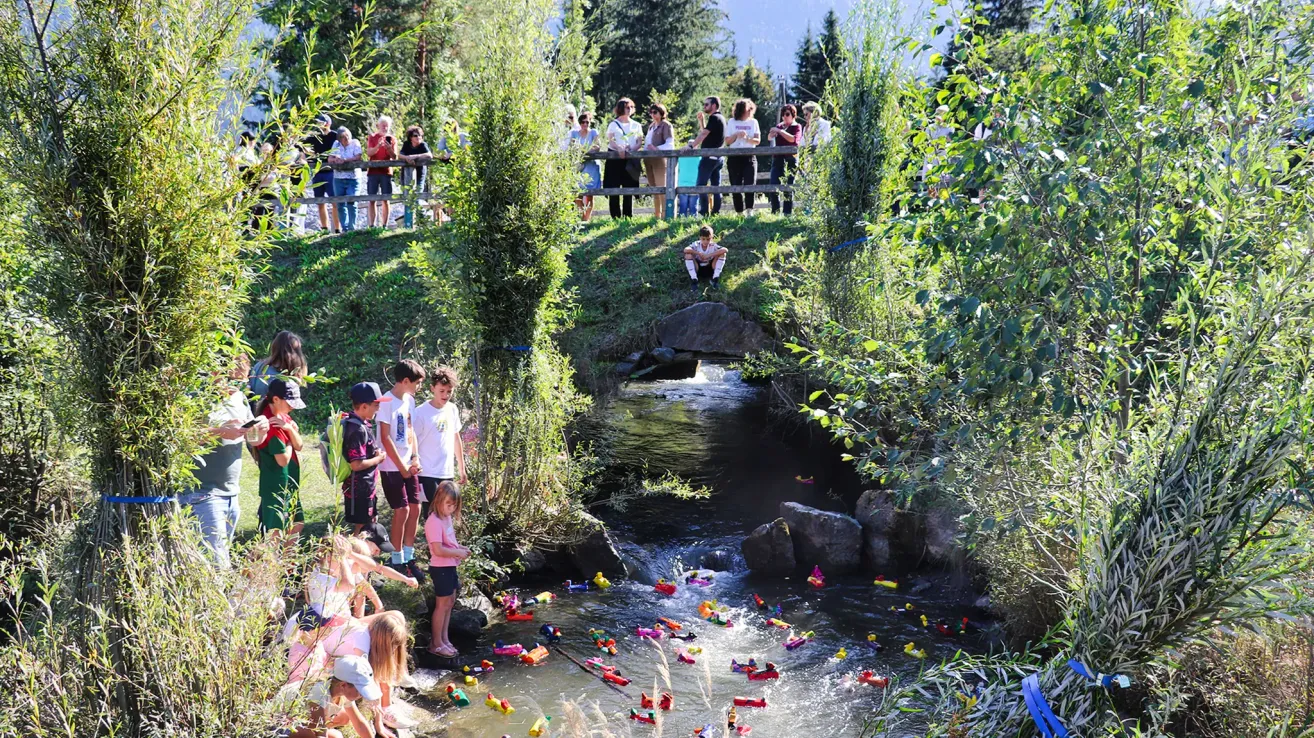 Eine Gruppe von Kindern und Erwachsenen beobachtet, wie Spielzeugboote einen kleinen Fluss hinuntertreiben. Einige sind auf einer Brücke, andere am Ufer.