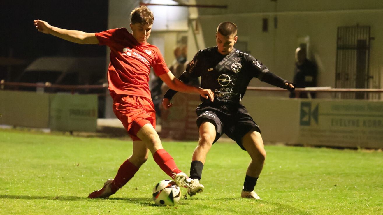Zwei Fußballer, einer in Rot und einer in Schwarz, kämpfen auf einem Feld bei Nacht um den Ball.