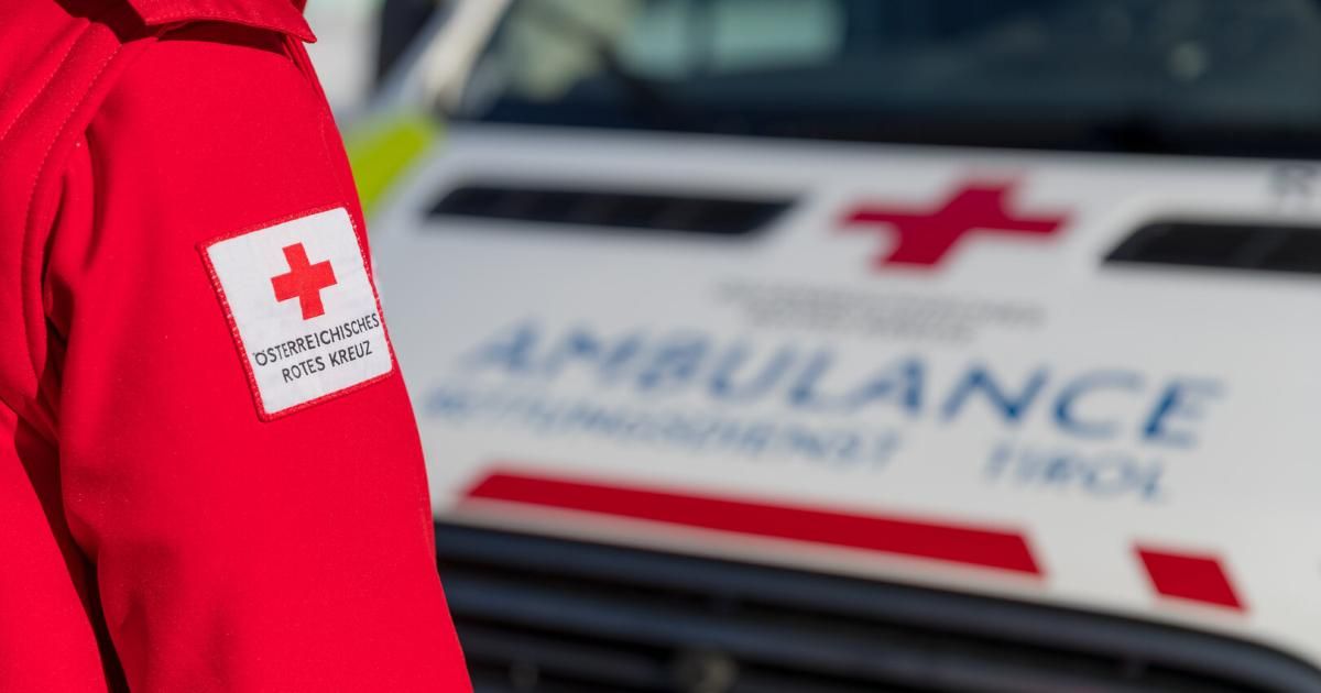 Nahaufnahme eines roten Aufklebers mit der Aufschrift 'Österreichisches Rotes Kreuz' auf einem roten Jackett, mit einem Krankenwagen im Hintergrund.
