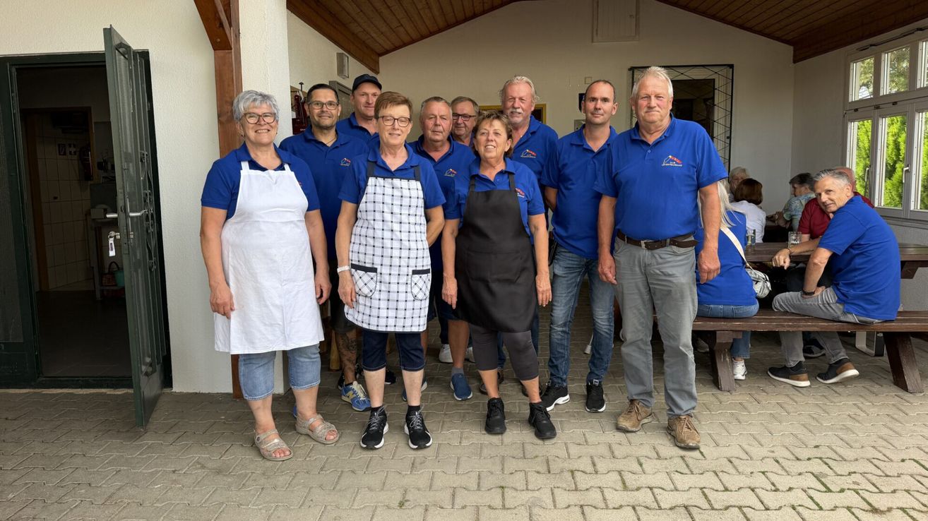 Eine Gruppe von Menschen in blauen Hemden und Schürzen steht zusammen in einem gut beleuchteten Raum. Einige tragen Brillen, und eine Person trägt ein Armband. Der Boden ist aus Ziegelsteinen, und hinter ihnen steht eine Holzbank.