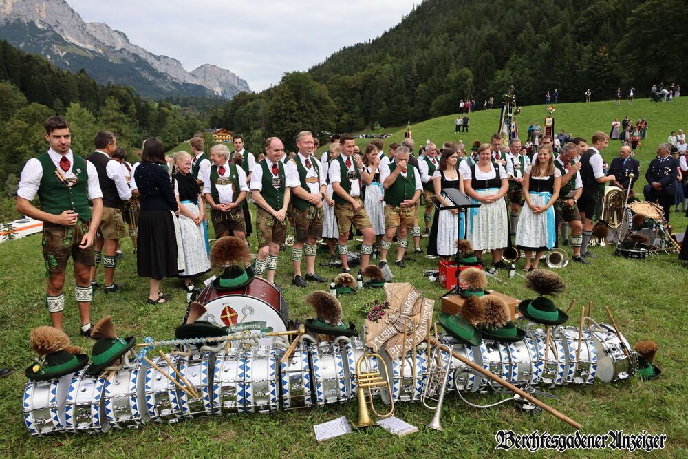 A group of people dressed in traditional Bavarian attire are standing in a grassy field with mountains in the background. Musical instruments are laid out in front of them.